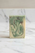 Load image into Gallery viewer, A rectangular bar of handmade soap with a green and cream marbled swirl pattern, resting on a gray wooden slatted surface outdoors.