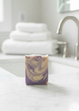 Load image into Gallery viewer, A rectangular bar of marbled soap with purple and cream swirls, standing on a white marble bathroom counter with a blurred sink, faucet, and folded towels in the background.
