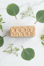 Load image into Gallery viewer, A rectangular bar of handmade soap with a honeycomb pattern, styled on a white marble surface with green eucalyptus leaves and small white flowers.