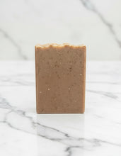 Load image into Gallery viewer, A rectangular bar of brown speckled soap or shampoo standing upright on a white marble surface with a matching marble background.