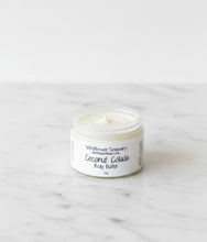 Load image into Gallery viewer, A small clear plastic jar of Wildflower Soaps Coconut Colada Body Butter sits on a white marble surface against a white background.