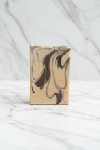 Load image into Gallery viewer, A rectangular bar of soap with a beige base and dark brown marbled swirls, resting on a gray slatted wooden surface with an outdoor garden background.