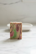 Load image into Gallery viewer, A rectangular bar of marbled soap with swirls of pink, green, and dark brown on a beige base, standing upright on a white marble surface with a small white dish blurred in the background.