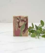 Load image into Gallery viewer, A rectangular handmade soap bar with a marbled swirl pattern of pink, green, beige, and dark brown, standing on a white marble surface next to green eucalyptus leaves.