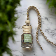 Load image into Gallery viewer, Herbal Indulgence car diffuser hanging from a decorative rope against a blurred green backdrop.