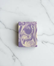 Load image into Gallery viewer, A rectangular bar of handmade soap with a purple and cream marbled swirl pattern, resting on a dark gray wooden slatted surface.