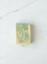 Load image into Gallery viewer, A rectangular bar of handmade soap with a marbled swirl pattern of light green and cream, resting on a gray slatted wooden surface.