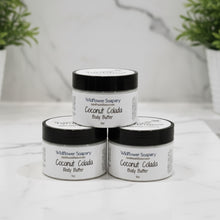 Load image into Gallery viewer, Three jars of Wildflower Soapary Coconut Colada Body Butter arranged in a pyramid on a white marble surface with blurred green plants in the background.
