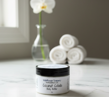 Load image into Gallery viewer, A jar of Wildflower Soapsy Coconut Colada Body Butter sits on a marble countertop in a bathroom setting, with rolled towels and a vase of an orchid blurred in the background.
