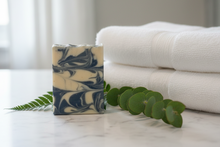 Load image into Gallery viewer, Sandalwood Goat Milk Soap on a marble countertop with white towels and greenery in the background for a serene spa experience.

