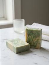 Load image into Gallery viewer, Two rectangular handmade soap bars with green and cream marbled swirls, displayed on a white marble surface with a blurred frosted glass and white towel in the background.
